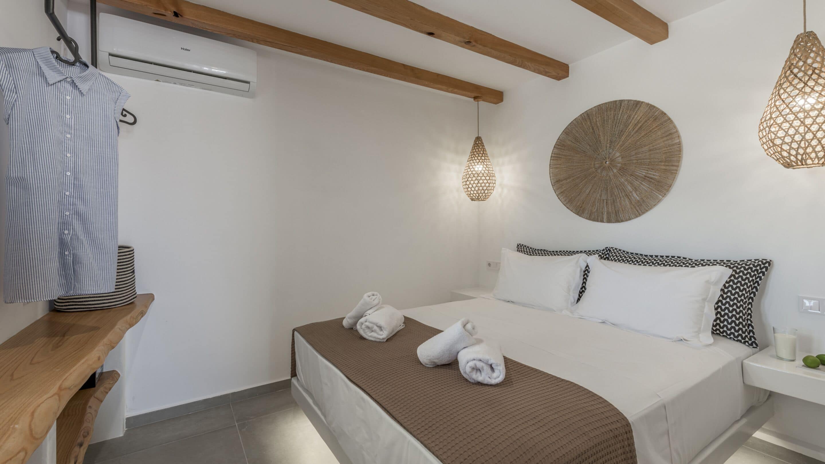 A hotel room with a bed, a wooden bench, a hanging light fixture, and a wall-mounted air conditioner.