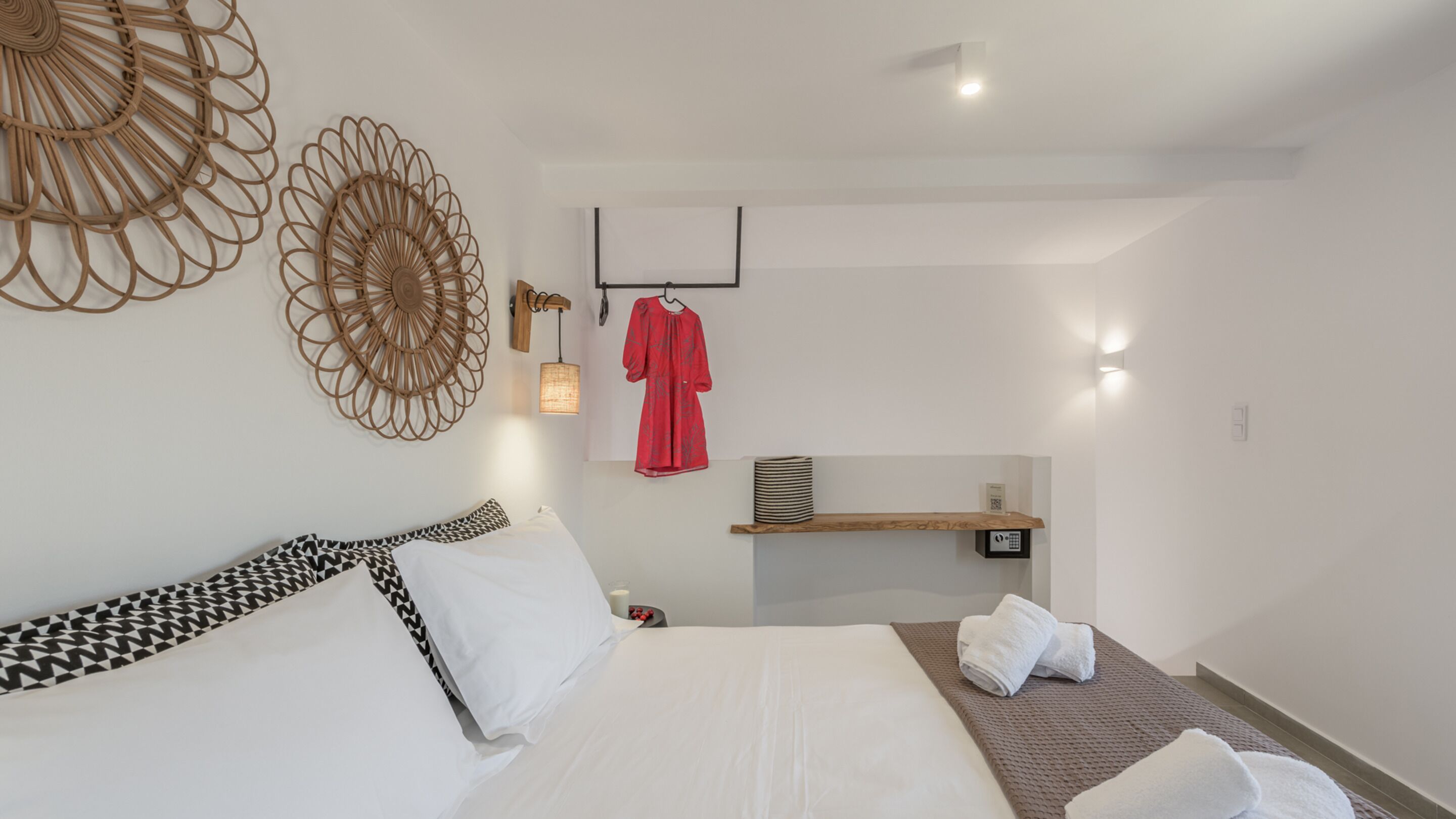 Une chambre d’hôtel moderne avec un grand lit, une étagère en bois où sont posées des serviettes, une robe rouge suspendue et des œuvres d’art décoratives accrochées au mur.