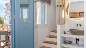 Un interior moderno con una puerta azul que da a un balcón, una escalera de madera y un espejo redondo sobre un lavamanos.