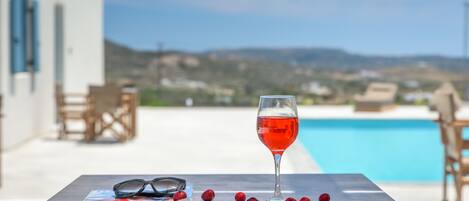 A table with a glass of red wine, glasses, and cherries, overlooking a pool and a scenic view.