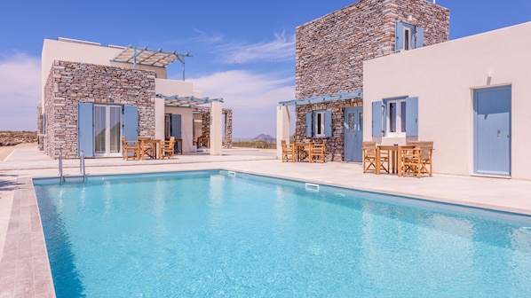 A pool area with stone buildings, outdoor seating, and a clear view of the sky. - Elements of Milos (Milos)