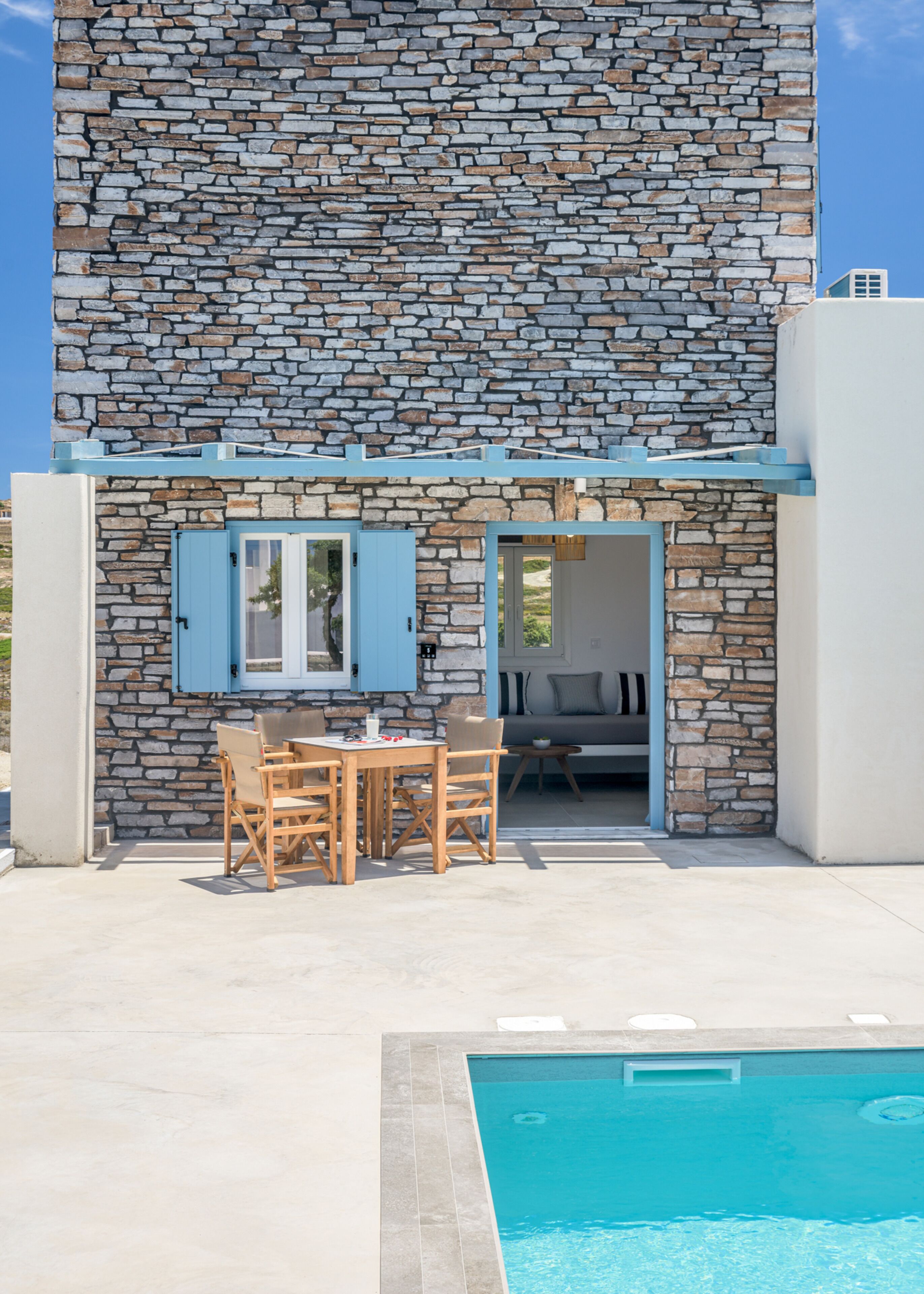 Un edificio de piedra con piscina, zona de comedor con sillas y mesa, y una puerta azul.