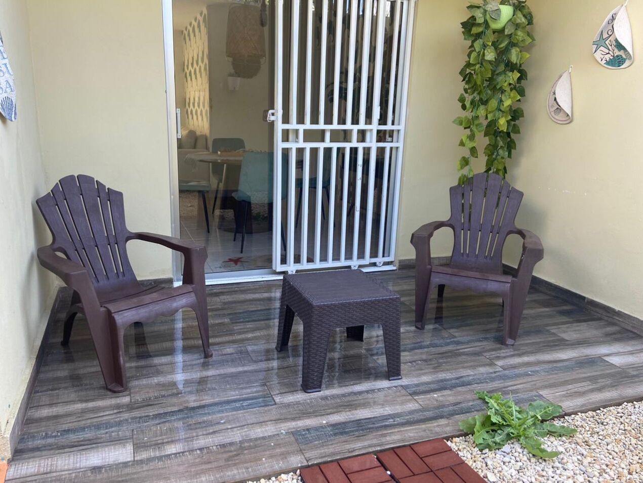 Een overdekt terras met twee stoelen en een kleine tafel, een wit hekwerk en een muur met decoratieve elementen.
