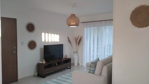 A living room with a TV on a wooden stand, a sofa, a decorative vase with plants, and a large window with sheer curtains. - Lovely condo with pool near beach (La Romana)