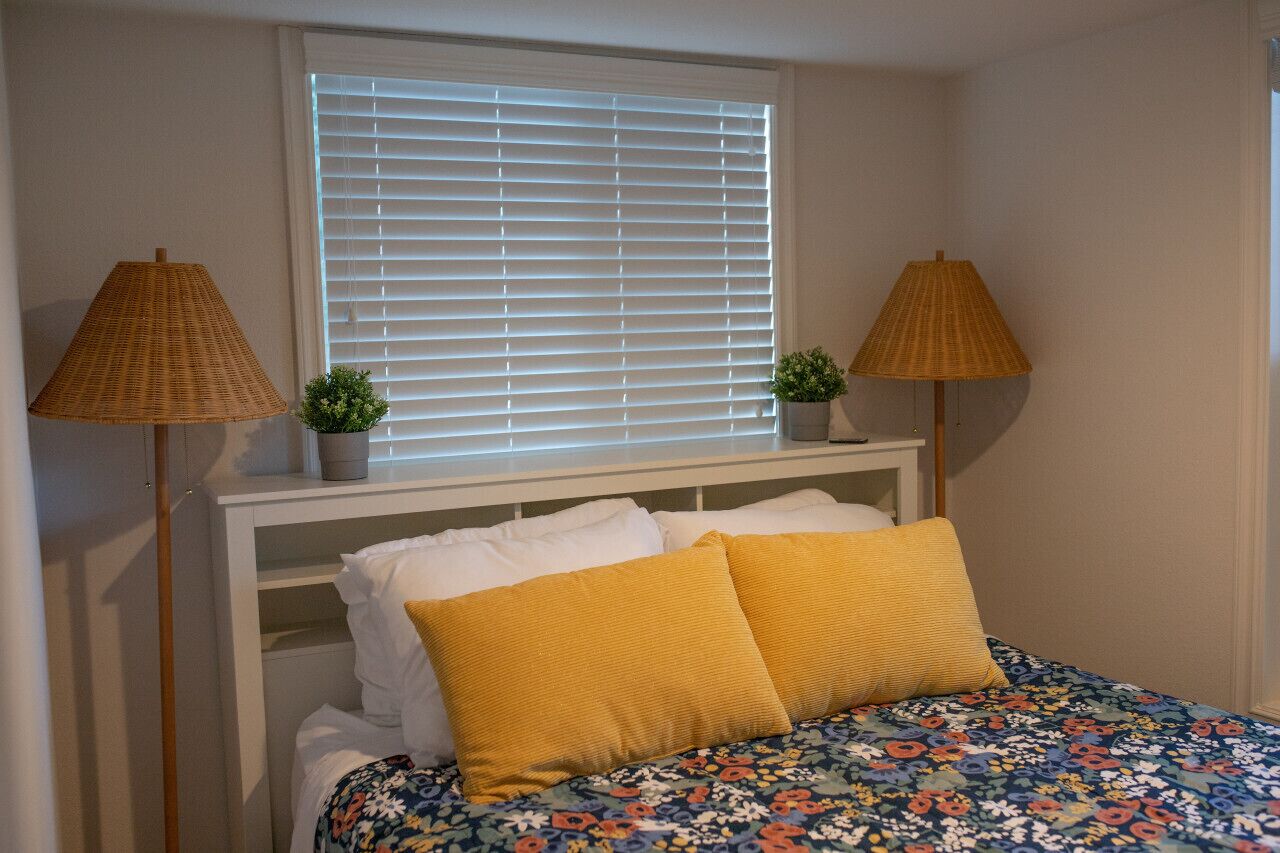 A bedroom with a bed, two lamps, a window with blinds, and a small plant on the nightstand.