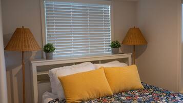 A bedroom with a bed, two lamps, a window with blinds, and a small plant on the nightstand.