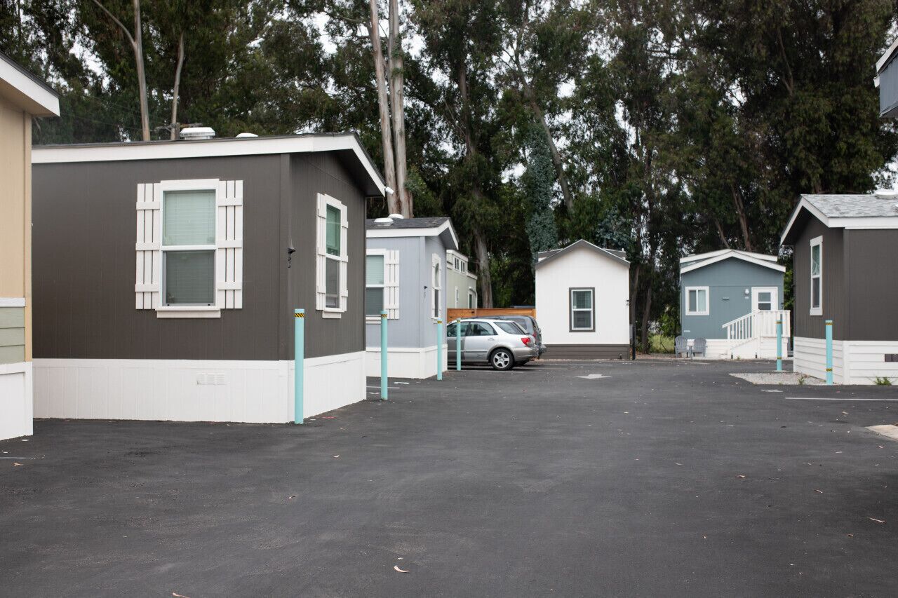 Uma fileira de pequenas cabanas térreas, com detalhes brancos e revestimento escuro, dispostas num estacionamento onde um carro está estacionado na frente.