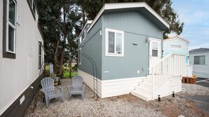 A small, single-story mobile home with a grey exterior and white trim. It features a front porch with a white railing and steps, two grey Adirondack chairs, and a small wooden bench.