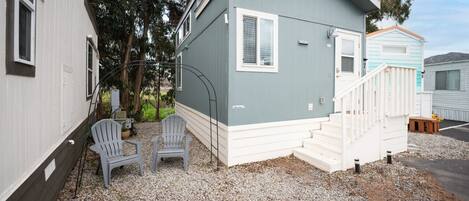 A small, single-story mobile home with a grey exterior and white trim. It features a front porch with a white railing and steps, two grey Adirondack chairs, and a small wooden bench.