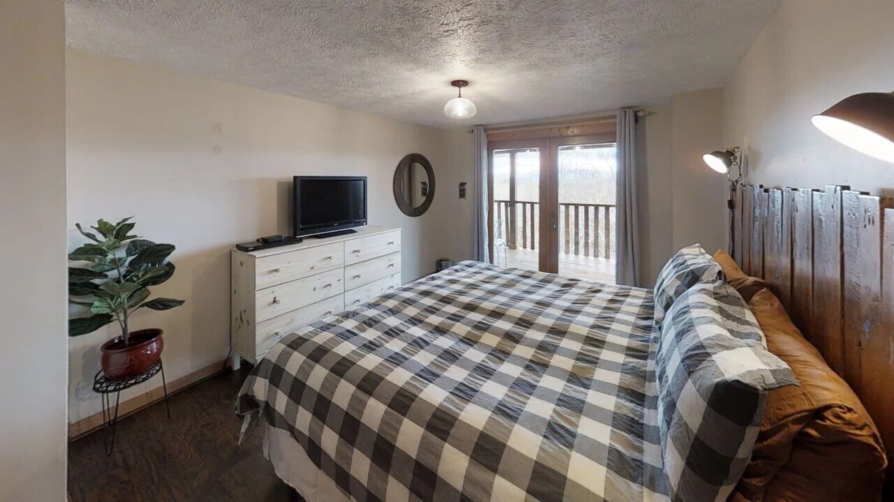 A bedroom with a bed, a television on a dresser, a potted plant, and a balcony with a view.