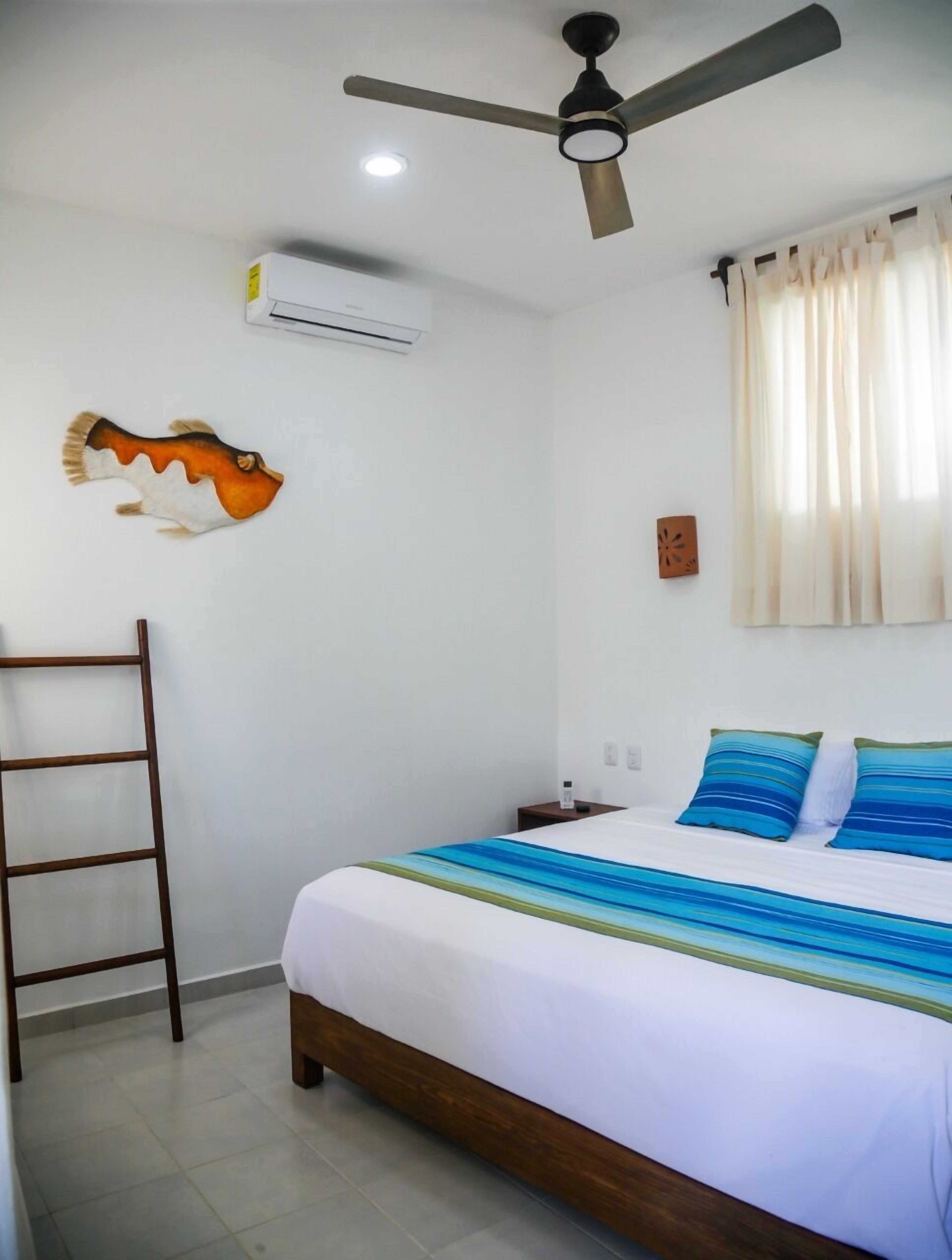 A hotel room with a bed, a ceiling fan, an air conditioner, a ladder, and a wall decoration.