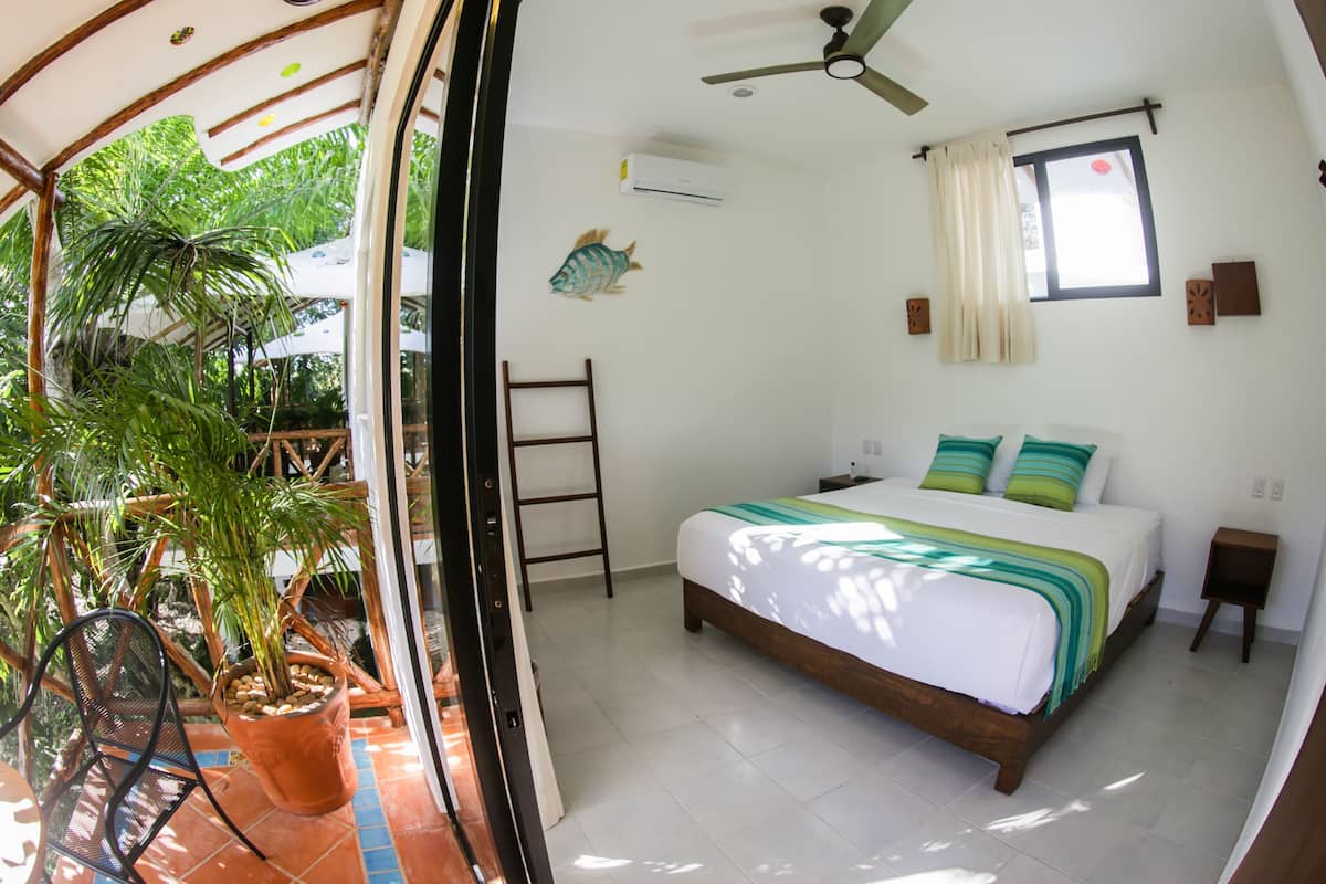 A bedroom with a bed, ceiling fan, air conditioning unit, window with curtains, and a potted plant.