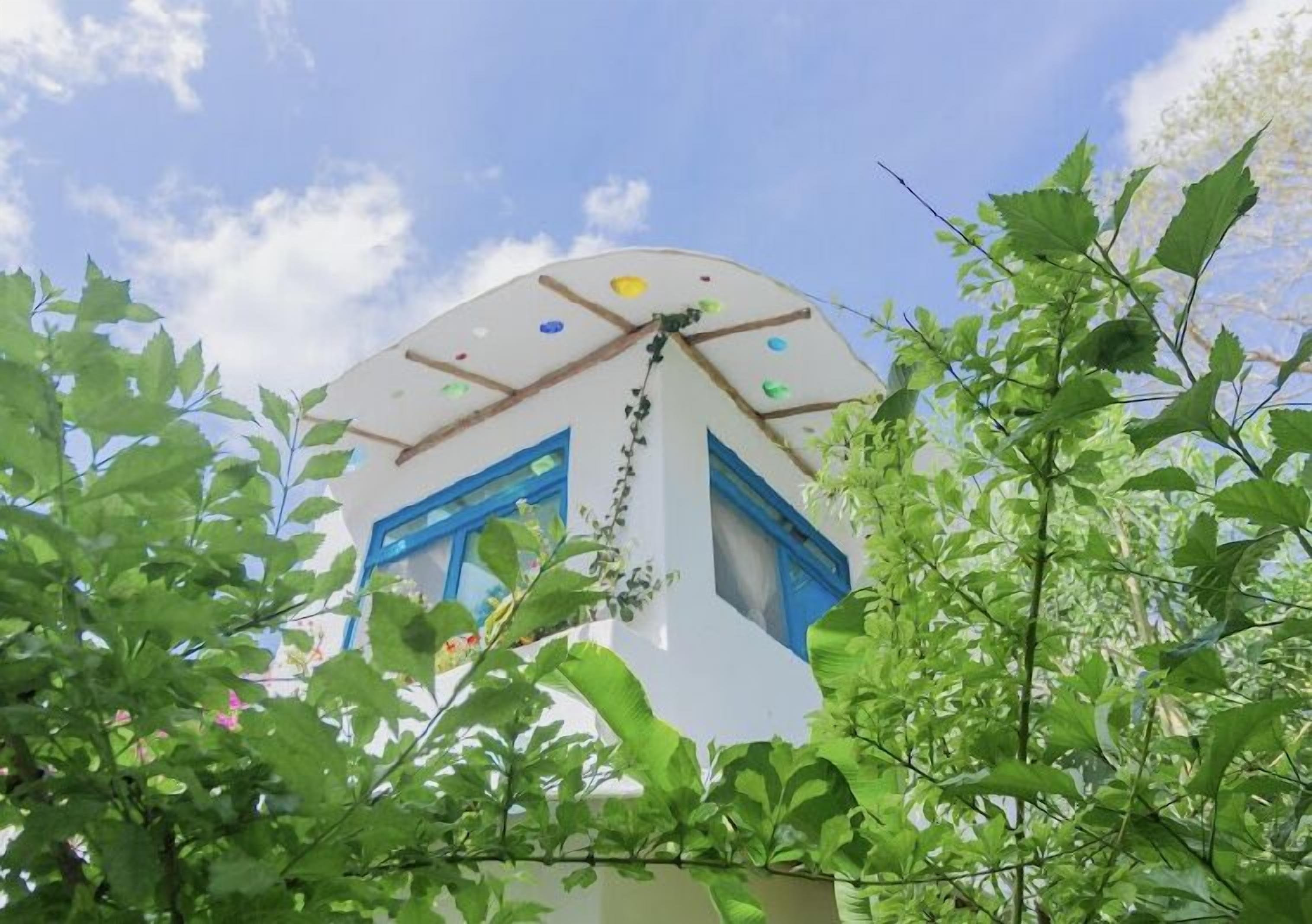 Un edifici amb façana blanca i finestres blaves, envoltat d'una vegetació verda abundant.