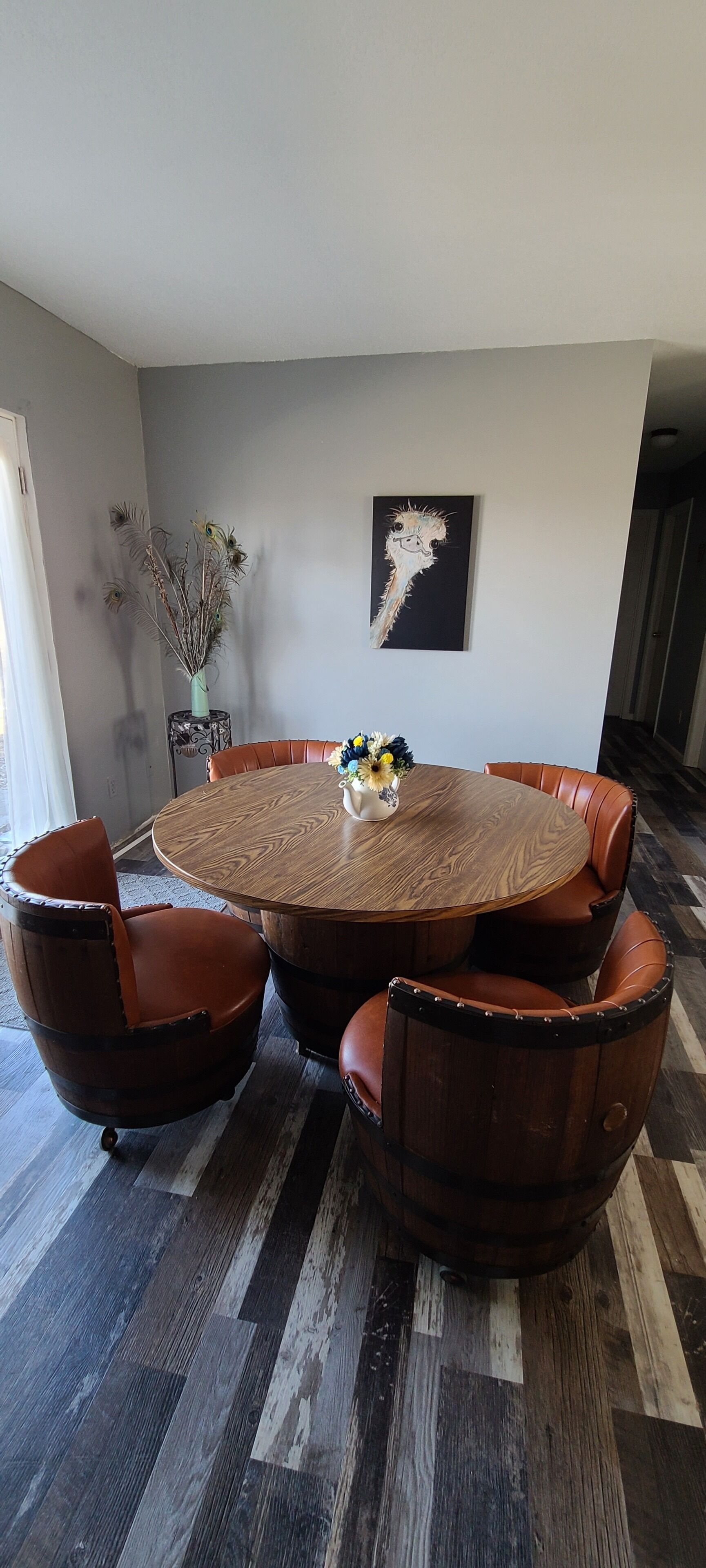 Une table de salle à manger ronde en bois, entourée de six chaises en cuir, un vase contenant des plantes séchées et un tableau accroché au mur.