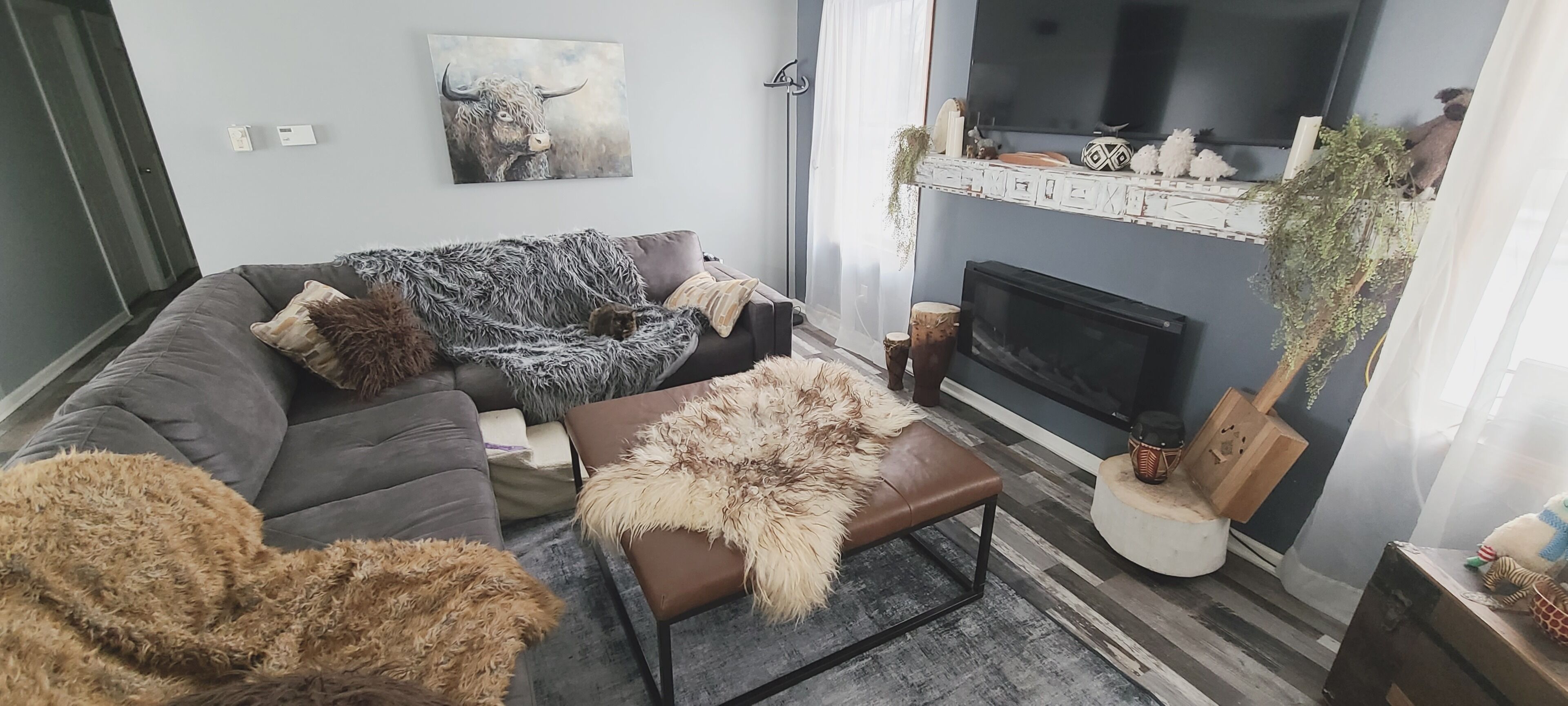 A living room with a grey sofa, a brown armchair, and a fluffy rug. There is a fireplace with a TV above it and a painting of a bull on the wall.
