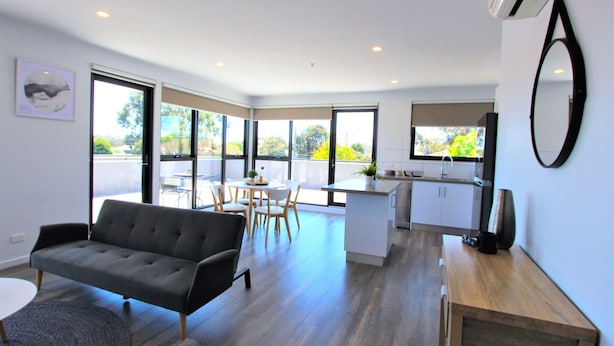 A modern apartment with a living room, dining area, and kitchen. It features large windows, a sofa, a dining table with chairs, and a kitchen counter. - StayCentral-Heidelberg Heights Penthouse (Heidelberg Heights)
