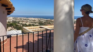 Ein Balkon mit Blick auf eine Küstenstadtlandschaft, eine weiße Säule und eine Person in einem weißen Kleid mit blauen Mustern.