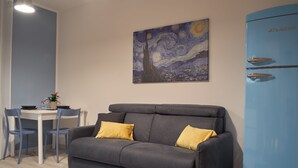 Un salon moderne avec un canapé gris, deux coussins jaunes, une table avec des chaises et un réfrigérateur bleu portant l’inscription « ATLANTIC ».