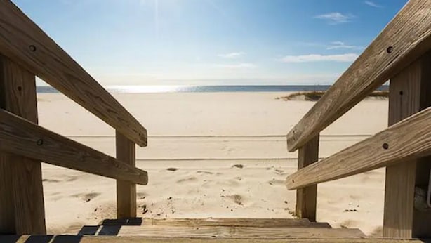 Ein hölzerner Strandpromenade führt zu einem Sandstrand unter blauem Himmel.