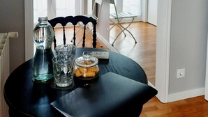 A dining area with a table set for two, featuring a glass bottle, a glass, and a jar of cookies. The room has a modern design with a dark-colored chair and a view of a hallway with a folding chair and a door. - Smiling Places (Vila do Conde)