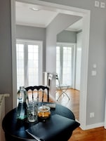 Un coin repas avec une table dressée pour deux personnes, agrémenté d’une bouteille en verre, d’un verre et d’un bocal de biscuits. La pièce présente un design moderne, avec une chaise de couleur foncée et une vue sur un couloir où l’on aperçoit une chaise pliante et une porte.