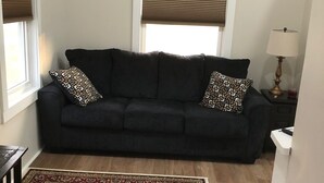 Uma sala de estar com um sofá azul escuro, almofadas decorativas estampadas, piso de madeira e um tapete ornamental.