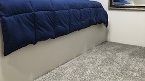 Quarto pequeno com cama de solteiro, teto inclinado, espelho grande na parede e piso coberto por carpete.