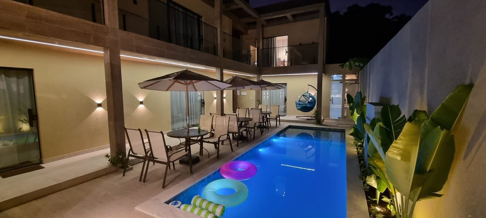 A swimming pool with lounge chairs and umbrellas, surrounded by a building with balconies and a glass wall.