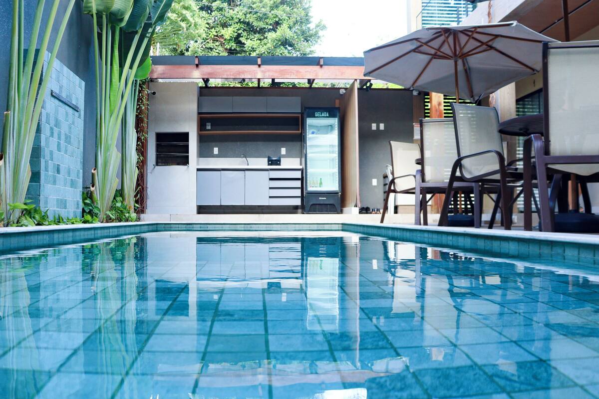 A swimming pool with tiled edges, a seating area with chairs and an umbrella, and a refrigerator labeled "GELADA" in the background.