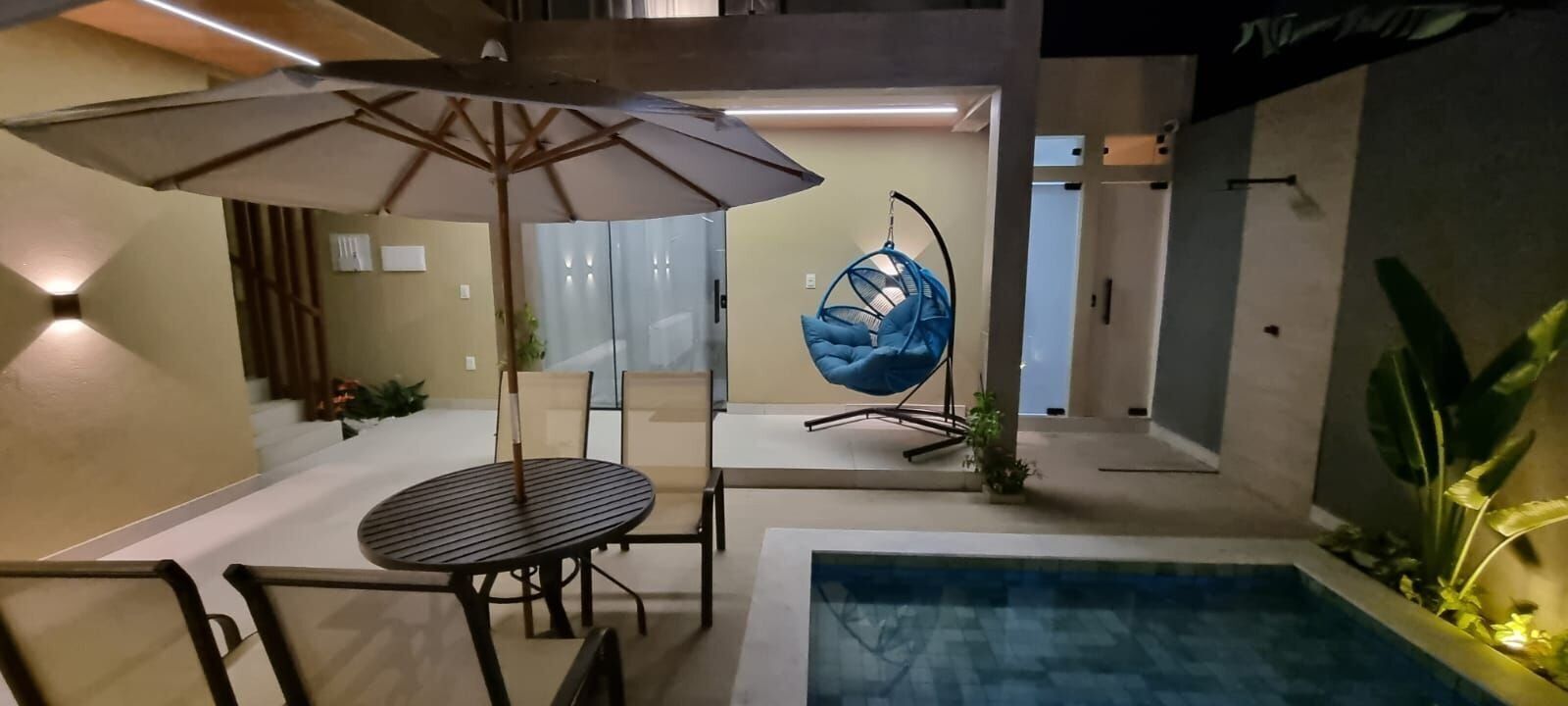 En moderne udendørs terrasse med pool, spisebord med stole, hængekøjsstol og stor parasol.