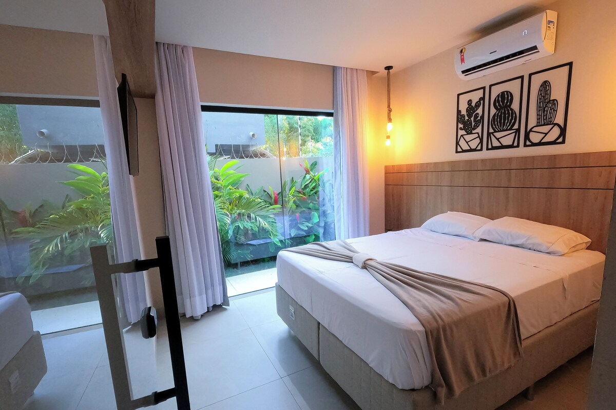A hotel room with a bed, a window with curtains, an air conditioning unit, and a wall with framed artwork.