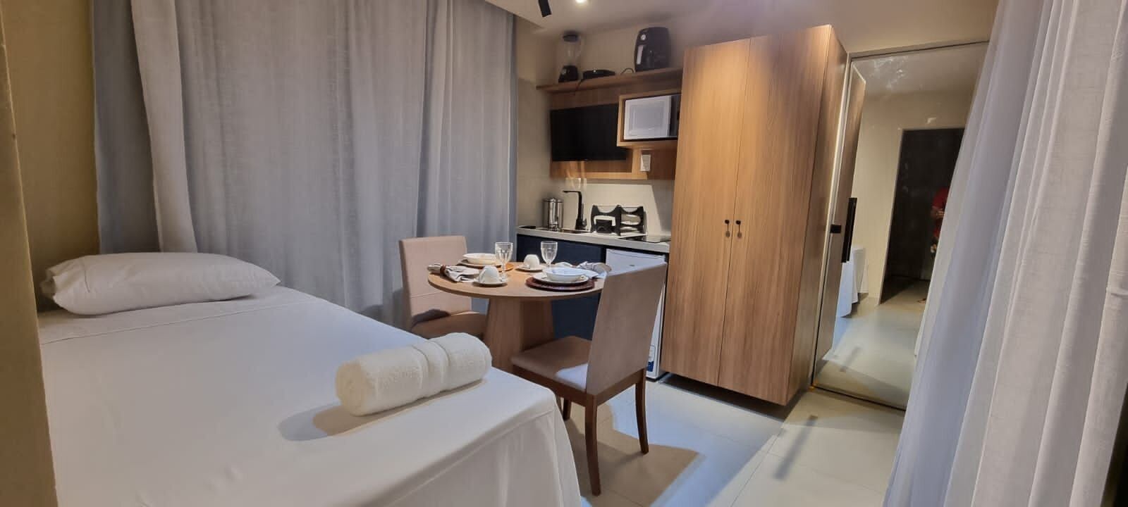 A compact hotel room with a bed, a small table with a chair, a kitchenette, and a door leading to another room.