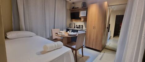 A compact hotel room with a bed, a small table with a chair, a kitchenette, and a door leading to another room.