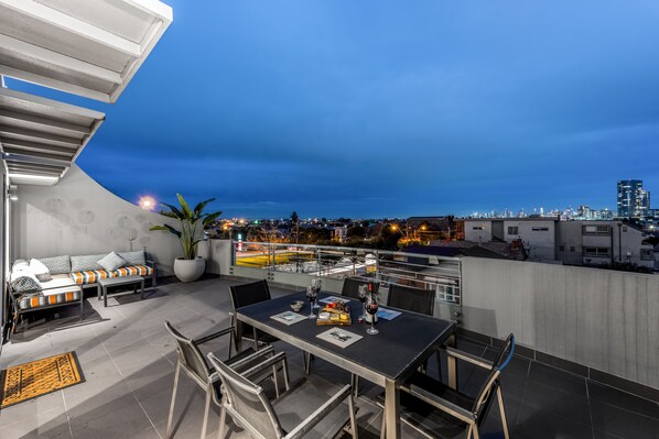 A rooftop terrace with a dining table, chairs, a sofa, and a potted plant.