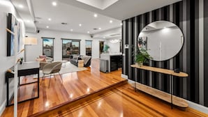 A modern living room with a wooden floor, a dining area with a table and chairs, a kitchen, and a large round mirror on the wall. - StayCentral - Moonee Ponds Penthouse (Moonee Ponds)