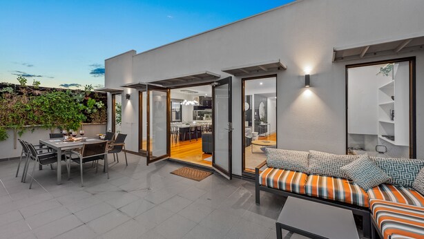 A modern outdoor patio with a dining area, a sofa with patterned cushions, and a kitchen visible through an open door. - StayCentral - Moonee Ponds Penthouse (Moonee Ponds)