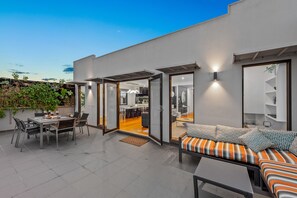 A modern outdoor patio with a dining area, a sofa with patterned cushions, and a kitchen visible through an open door. - StayCentral - Moonee Ponds Penthouse (Moonee Ponds)