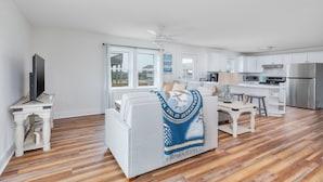 Een ruime woonkamer met een witte bank, een televisie op een houten dressoir en een eethoek met een tafel en stoelen. De keuken is zichtbaar, met witte kastjes en roestvrijstalen apparatuur.