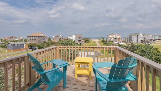 Een houten terras met turquoise stoelen en een gele tafel, met uitzicht op een strand en een woongebied.