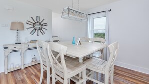 Een eetgedeelte met een witte houten tafel en stoelen, een decoratief wandobject, een lamp en een raam met gordijnen.