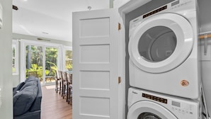 Ein Waschraum mit einer Whirlpool Waschmaschine und einem Trockner, ein graues Sofa und ein Essbereich mit Blick auf Bäume und eine Terrasse.