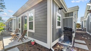 A single-story mobile home with a gray exterior, a front porch with two wooden chairs, a grill, and a small potted plant. - 1385 - Stay Awhile - Cozy Corolla Getaway Near Beach & Community Pool (Corolla)