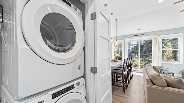 A laundry room with stacked washers and dryers, a dining area with a table and chairs, and a view of the outdoors through large windows. - 1385 - Stay Awhile - Cozy Corolla Getaway Near Beach & Community Pool (Corolla)