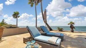 Eine Strandlounge mit zwei Liegestühlen, einem kleinen Tisch mit einer Serviertablett und Blick auf das Meer und Palmen.