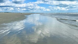 Une vaste plage aux vagues douces, un ciel dégagé et quelques personnes visibles au loin.