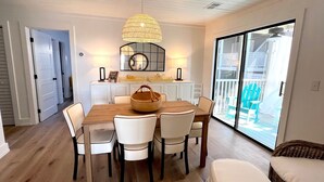 Un coin repas avec une table en bois, des chaises blanches, un panier en osier et une grande fenêtre donnant sur un balcon.