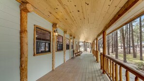 A covered wooden deck with a railing, windows, and a view of the surrounding forest. - Incredible Rustic Lakeside Cabin in the Woods! Near Lakes, Skiing and Trails! (Lakeside)