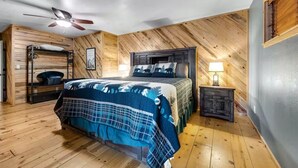 A bedroom with a bunk bed, wooden walls, a ceiling fan, a bedside table with a lamp, and a framed picture on the wall. - Incredible Rustic Lakeside Cabin in the Woods! Near Lakes, Skiing and Trails! (Lakeside)