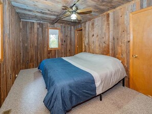 A bedroom with a bed, wooden walls, a ceiling fan, and a door.