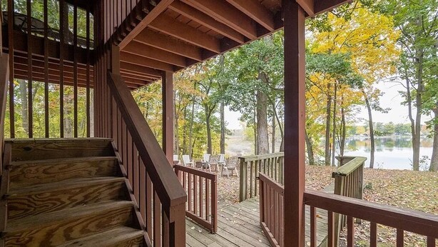 Eine Holztreppe führt zu einer überdachten Terrasse mit Blick auf einen See und Herbstlaub.